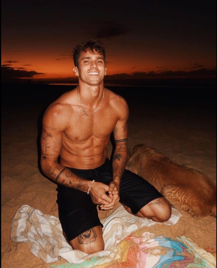 a shirtless man sitting on the beach next to his dog at sunset with an orange sky in the background