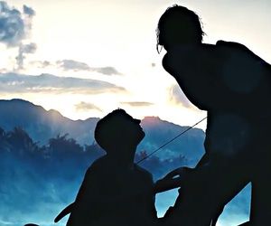 the silhouette of two men are standing in front of a mountain range with clouds and mountains behind them