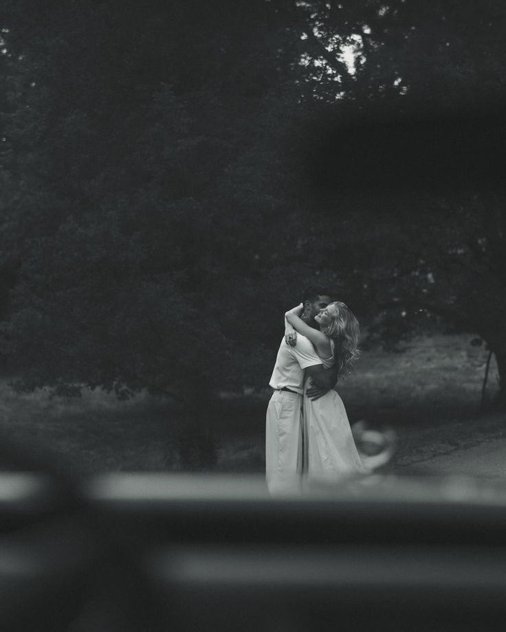 black and white photo of two people embracing each other in front of some trees on a road