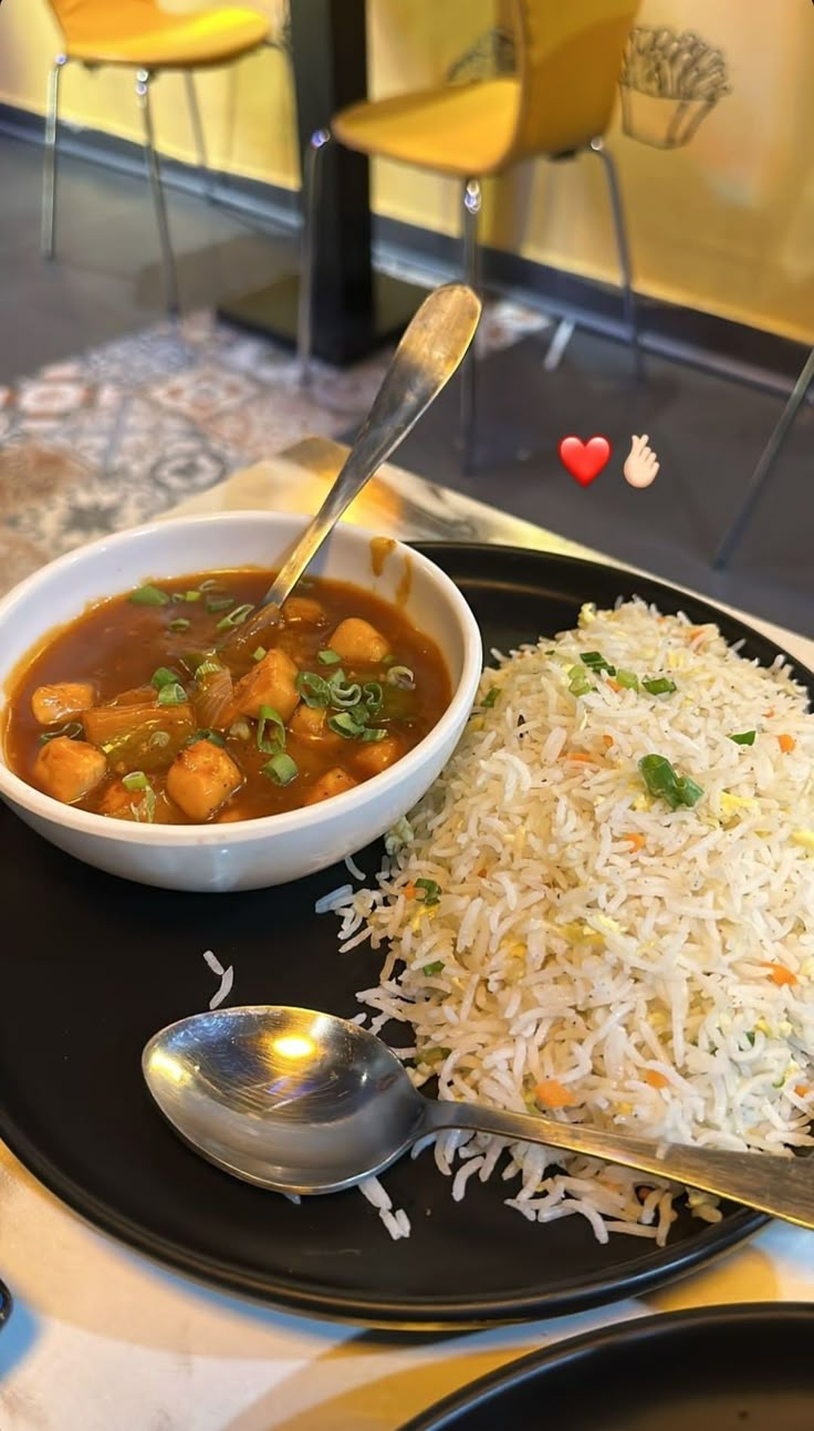 a black plate topped with rice and beans next to a bowl of soup on top of a table