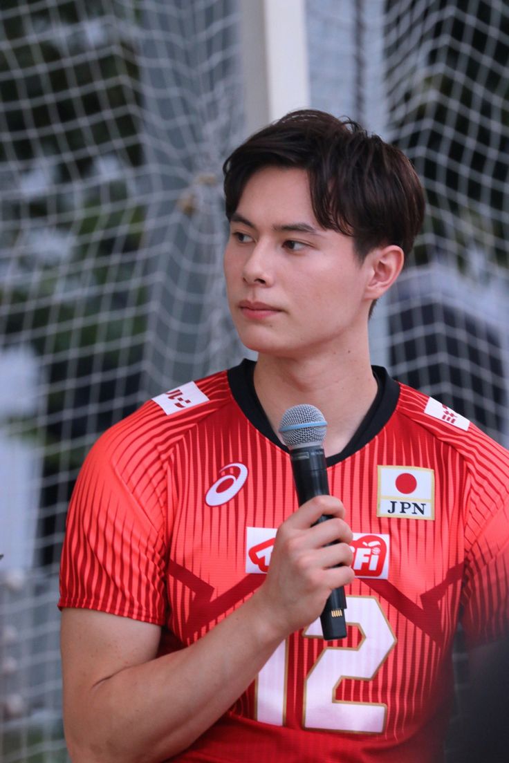 a young man holding a microphone in front of a net while wearing a red jersey