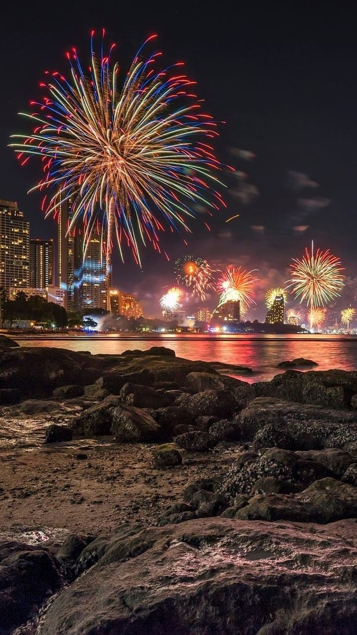 fireworks are lit up in the night sky over water and rocks on the beach with buildings in the background