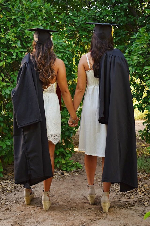 two women in graduation gowns holding hands and walking down a path with trees behind them