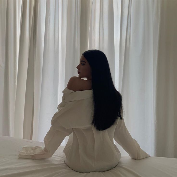 a woman sitting on top of a bed in front of curtains with her long black hair