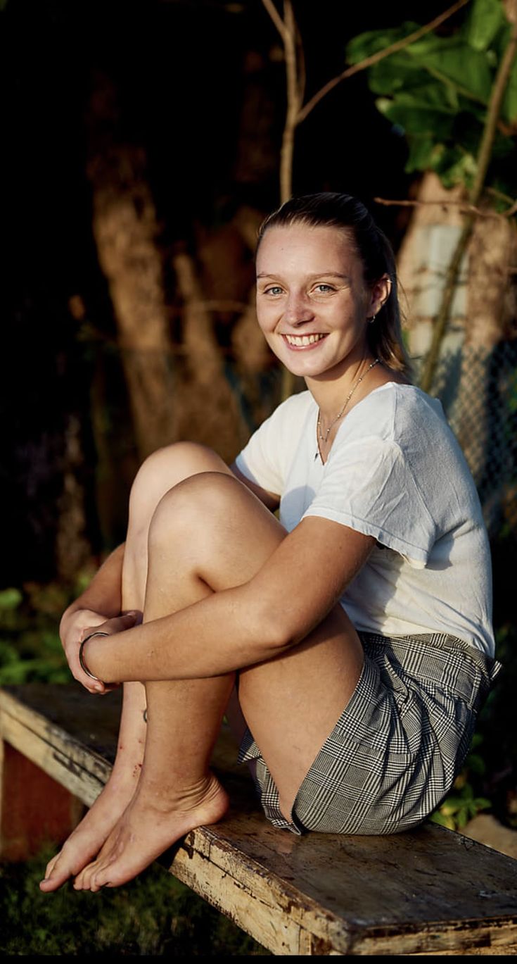 a young woman sitting on top of a wooden bench in front of some trees and bushes