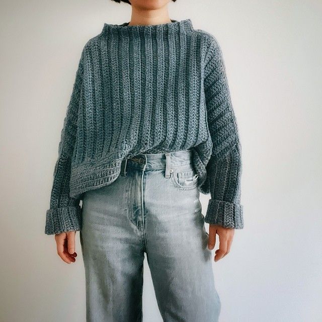 a woman standing in front of a white wall with her hands on her hips wearing jeans and a sweater