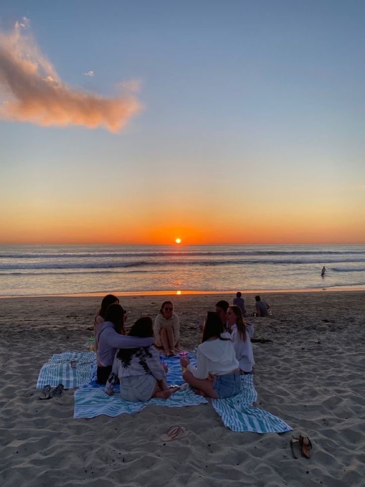 four people sitting on a blanket at the beach watching the sun set over the ocean