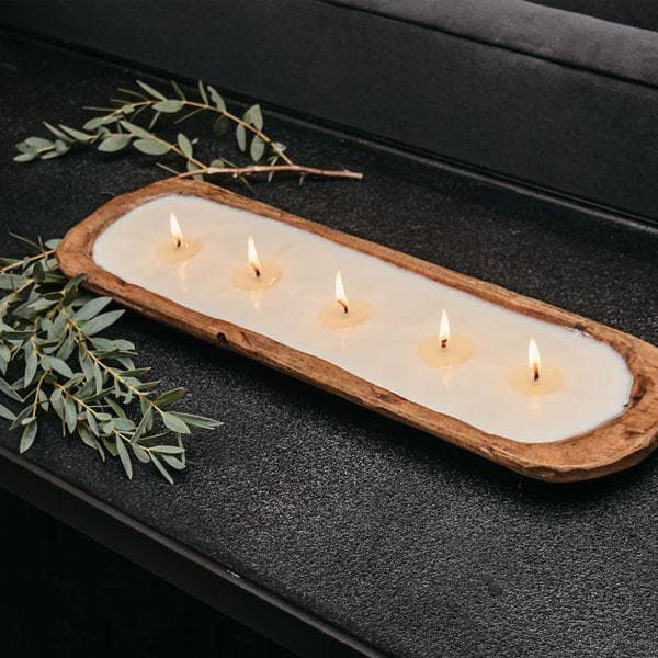 four lit candles in a wooden tray on a black surface with greenery around it