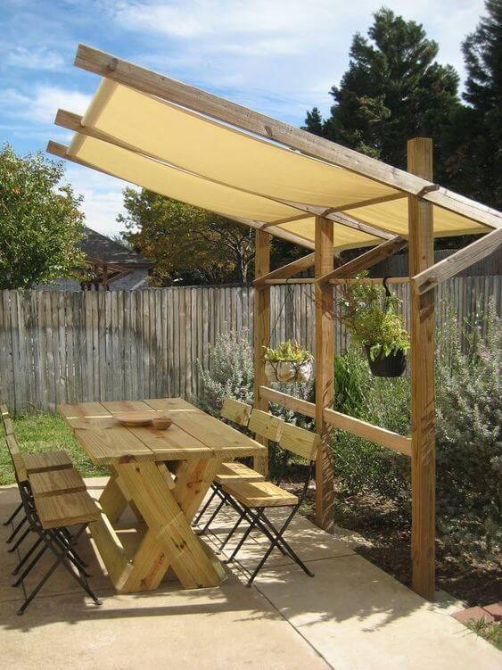 a wooden table and chairs under an awning next to a fence with trees in the background