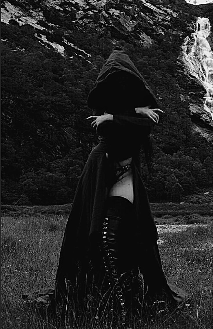 a woman in a black dress and hood with her hands on her face, standing in the grass