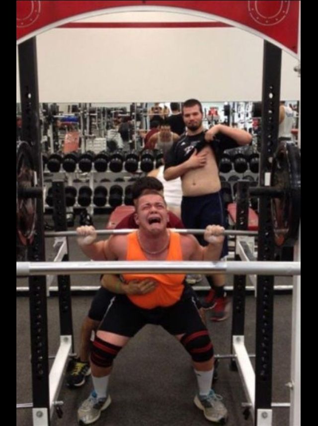 a man is squatting in the middle of a gym with another man behind him