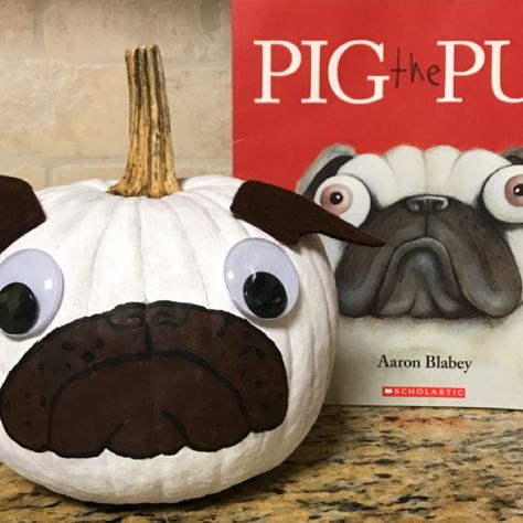 a white pumpkin with a dog's face painted on it next to a book about pug the pug