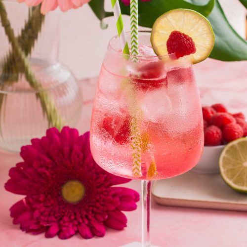 a pink cocktail with raspberries and lemon wedges on a table next to flowers