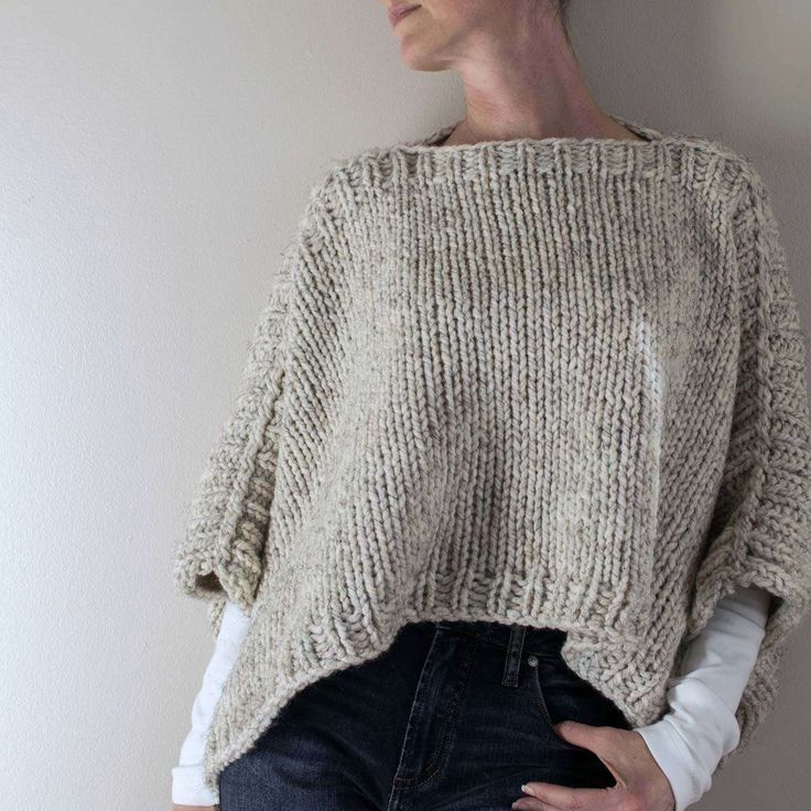 a woman standing in front of a white wall wearing a sweater and jeans with her hands on her hips