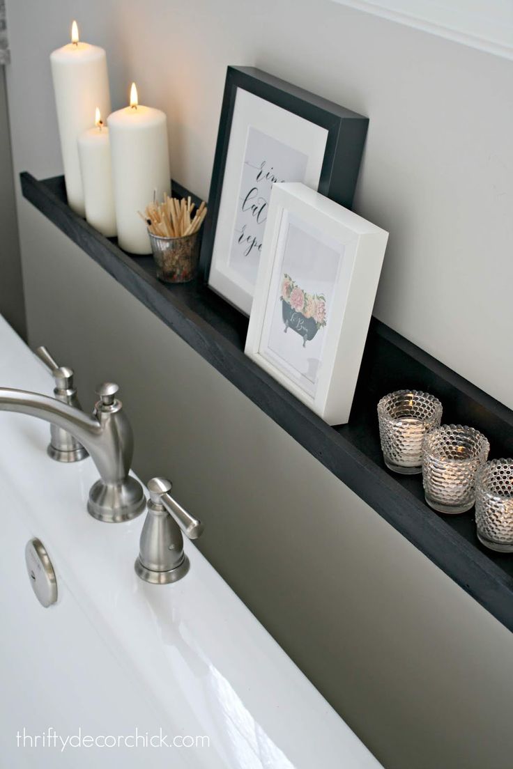 some candles are sitting on a shelf above a bathtub with two pictures and three votives