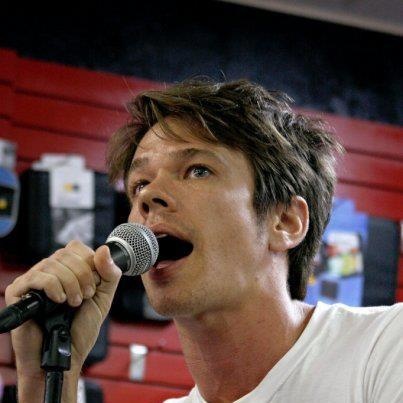 a man singing into a microphone in front of a wall full of red and black chairs