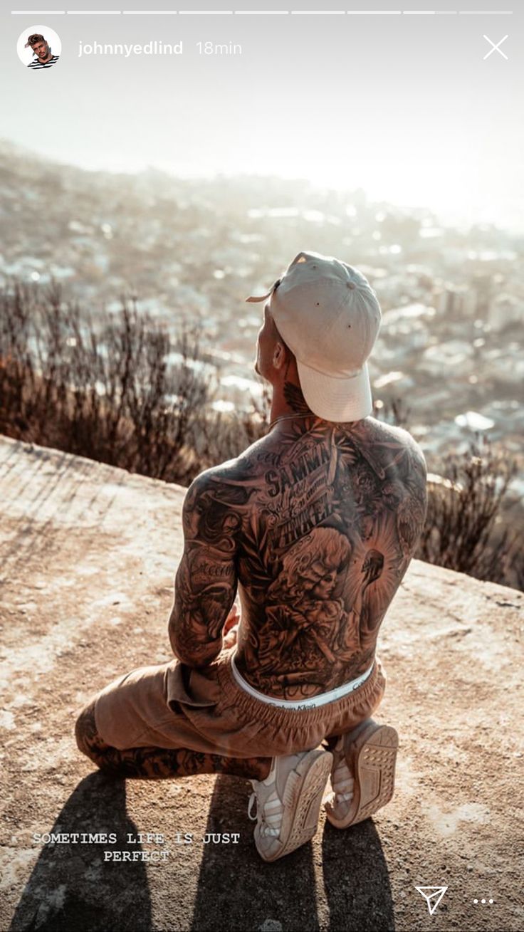 a man with tattoos sitting on top of a cliff looking out at the ocean and mountains