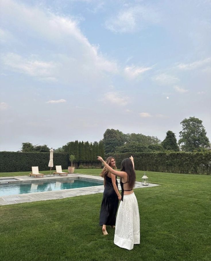 two women are standing in the grass near a pool and some trees with their arms around each other