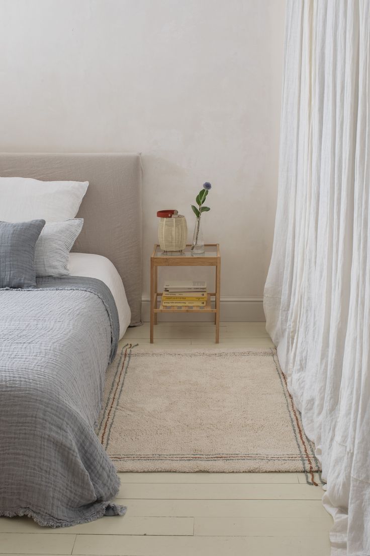 a bed sitting next to a window in a room with white curtains on the windowsill