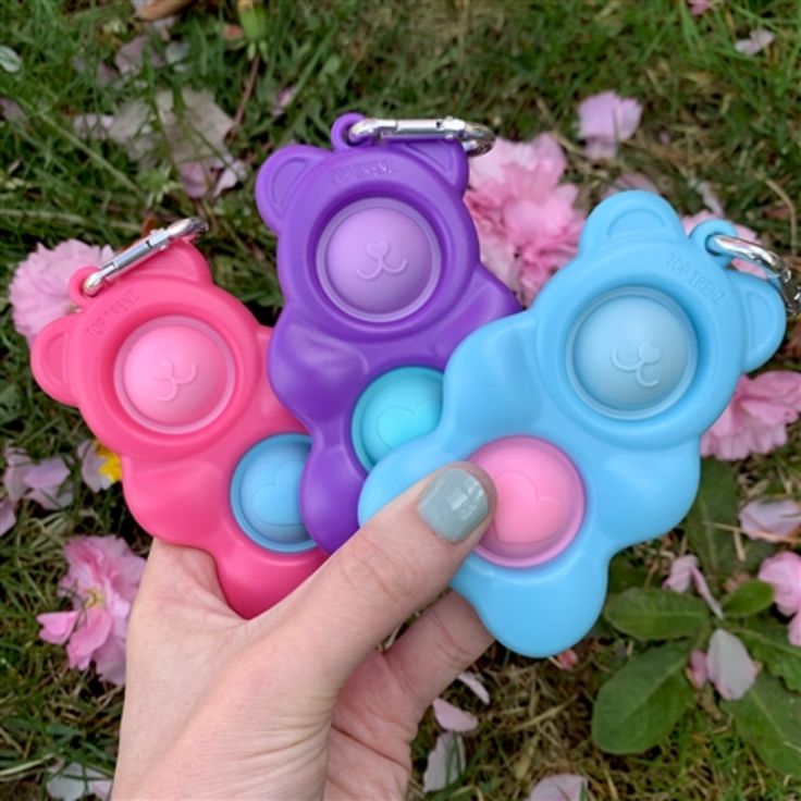 a hand holding three different colored teddy bears in front of some pink and blue flowers