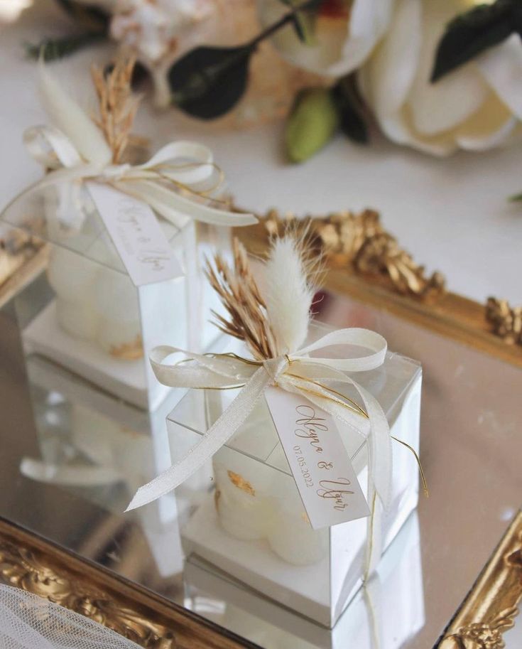 two clear boxes with white flowers and ribbon tied around them on a mirrored surface in front of a gold framed mirror