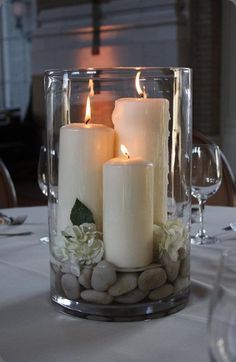 two white candles are sitting in a glass vase with rocks and greenery on the table