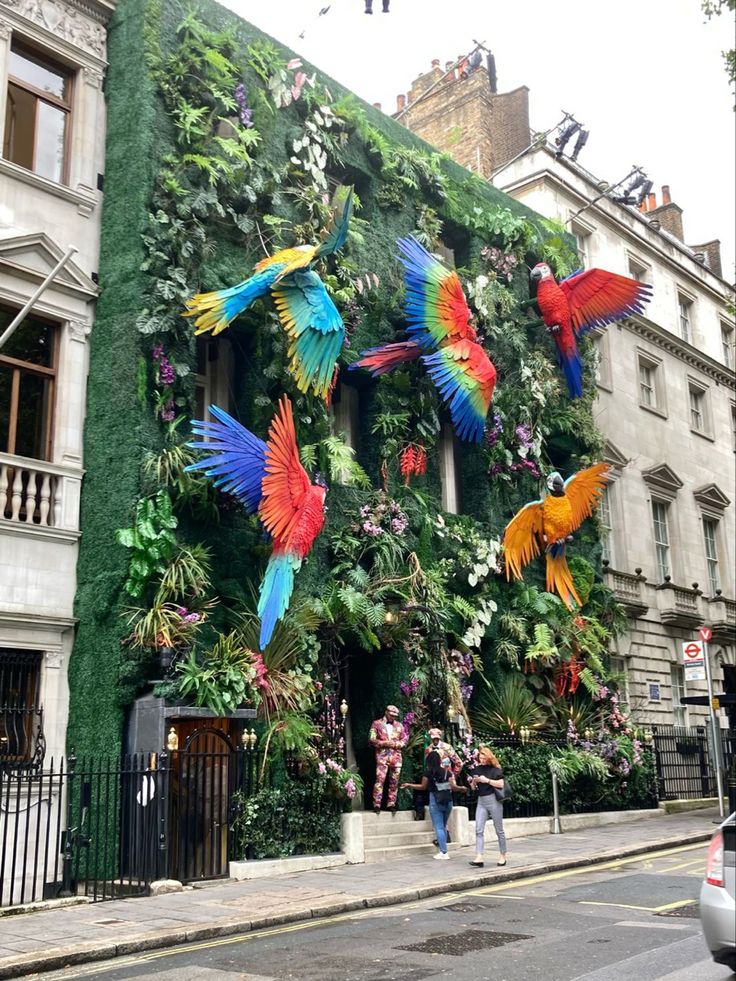 the building is decorated with colorful birds and plants on it's side, while people are walking down the street