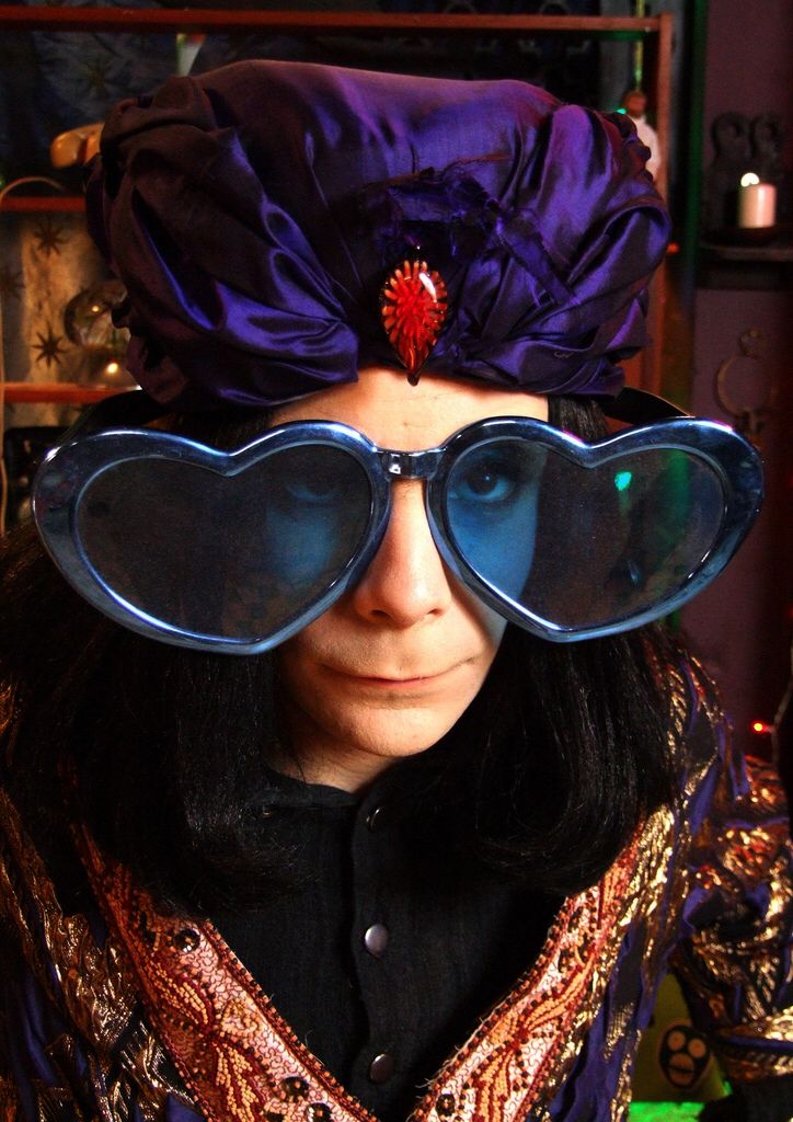 a woman wearing large blue glasses and a purple flower on top of her head is looking at the camera