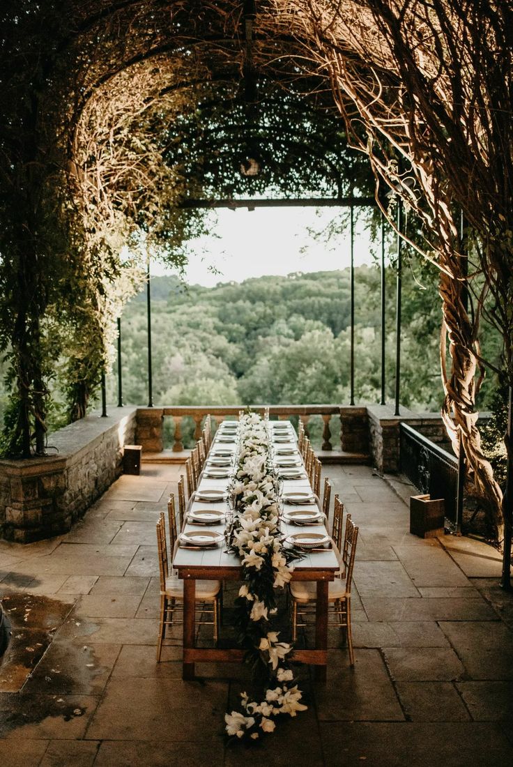 an outdoor dining table set up with white flowers and greenery on the tables, surrounded by vines