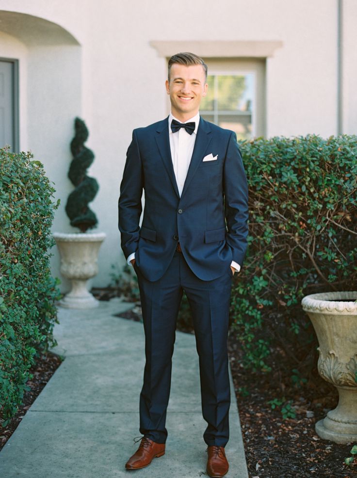 a man in a suit and bow tie standing on a sidewalk next to some bushes