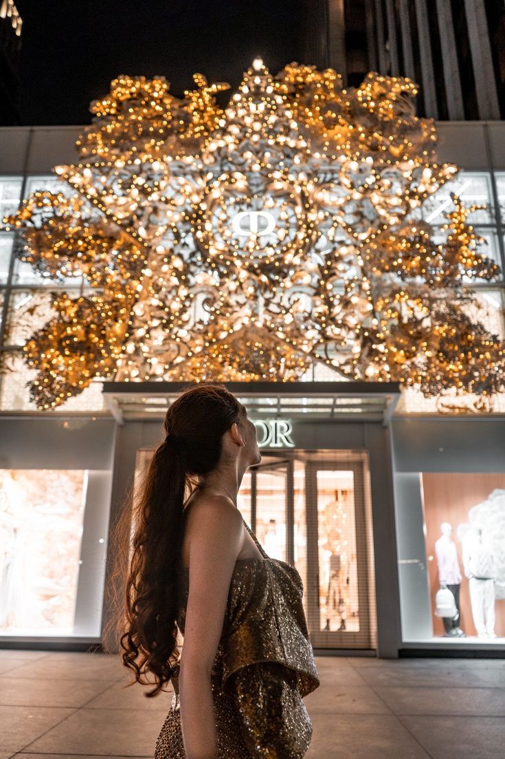 a woman standing in front of a building with lights on it's windows and trees behind her