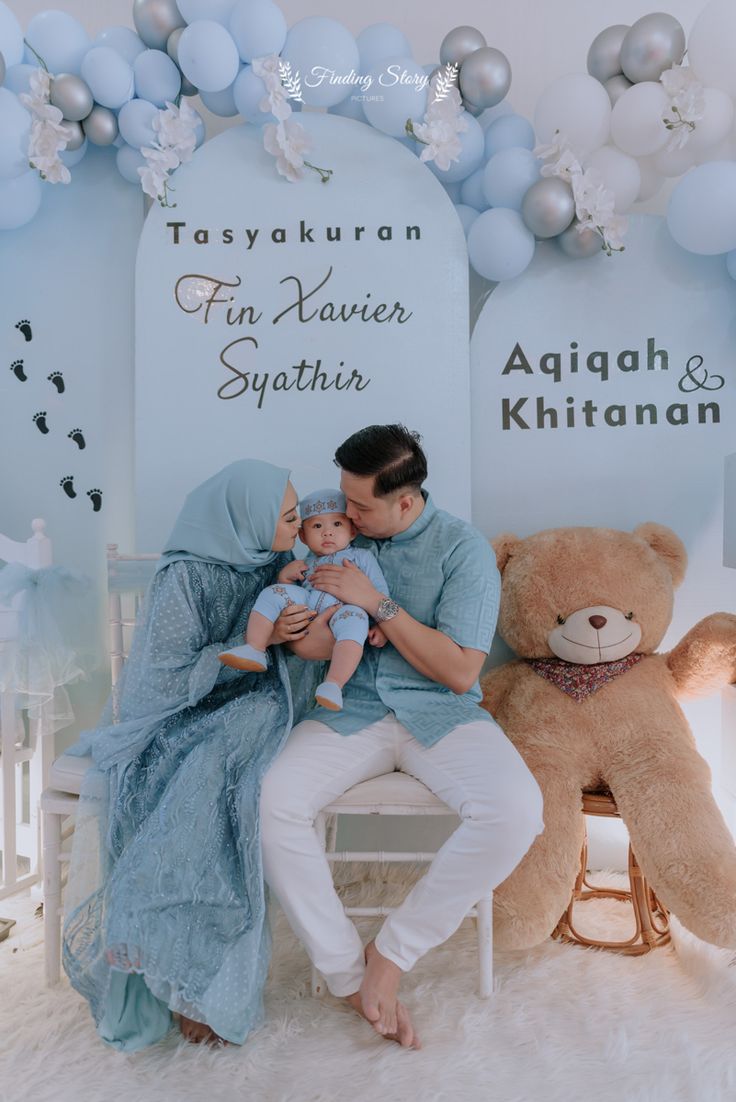 a man holding a baby while sitting in front of a teddy bear and balloon arch