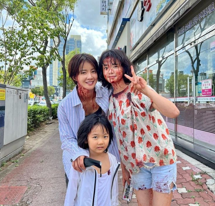 two women and a child with blood on their faces posing for a photo in front of a building