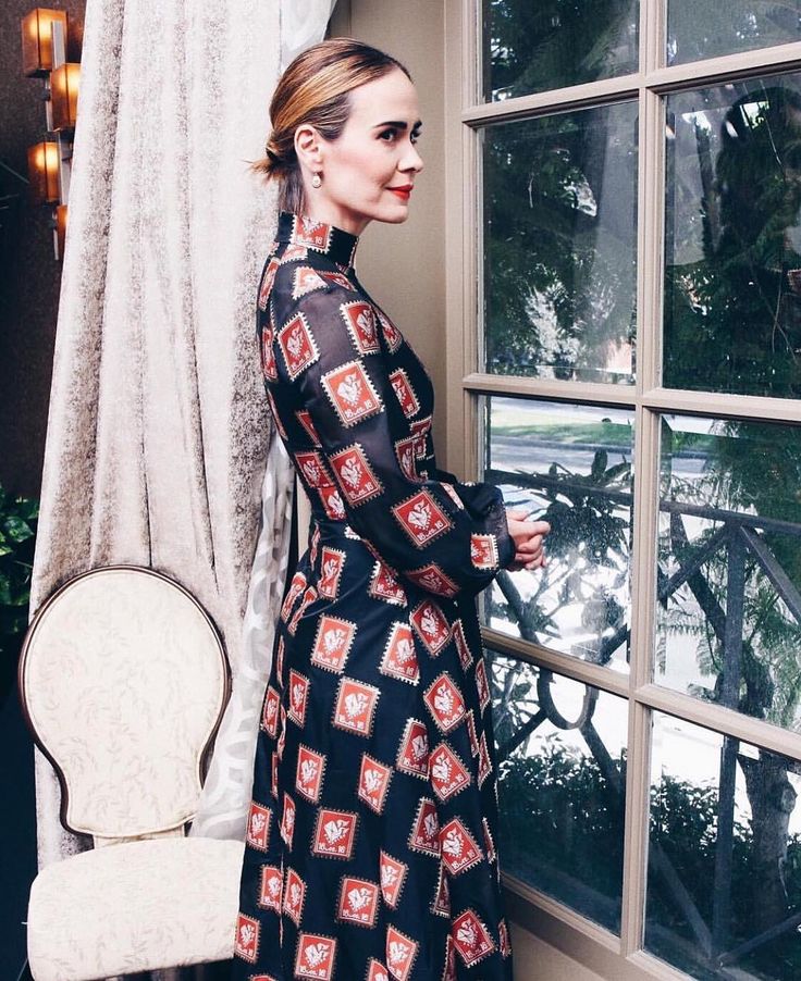 a woman in a black and red dress standing by a window with a white chair