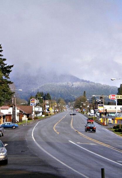 cars are driving down the street in front of buildings and mountains on a cloudy day