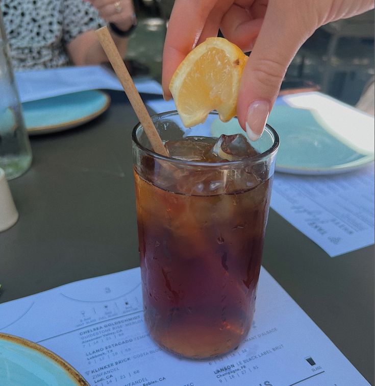 a person holding a lemon wedge over a glass filled with ice and soda on top of a table