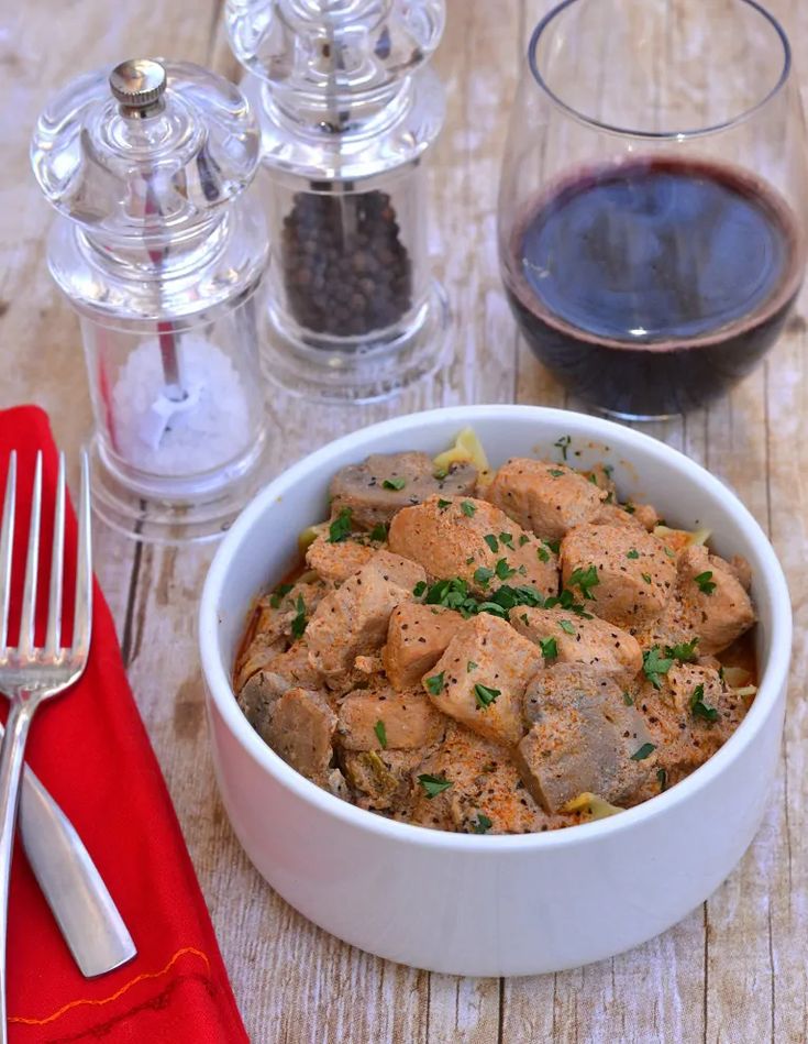 a white bowl filled with food next to a fork and glass of wine on top of a wooden table