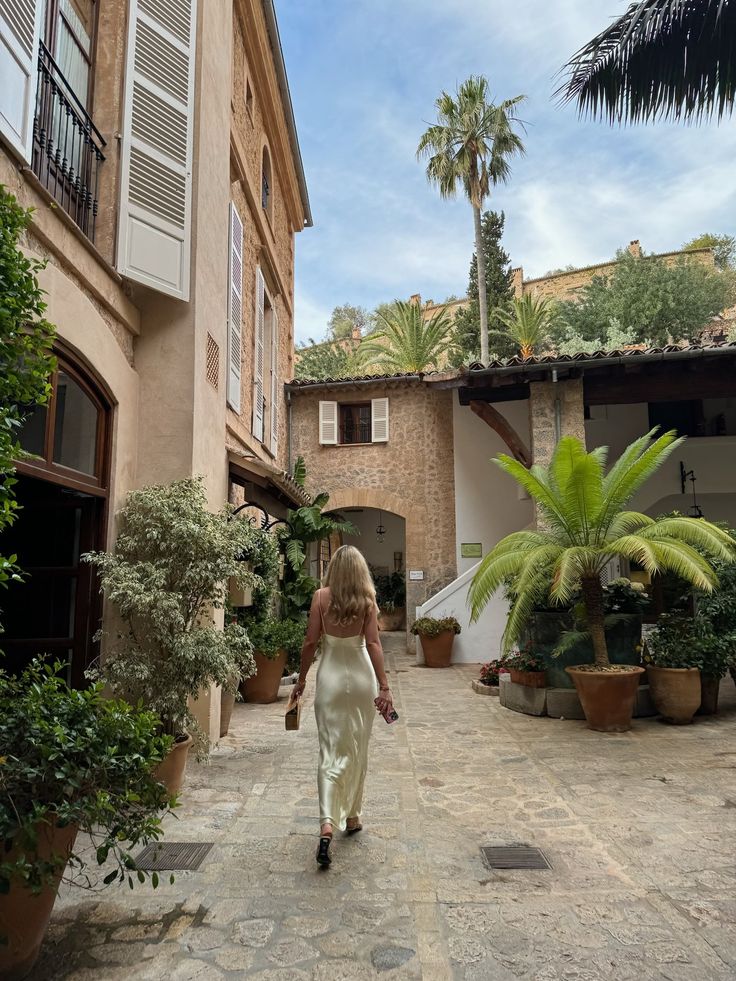 a woman walking down a street next to potted plants and palm trees on either side of the building