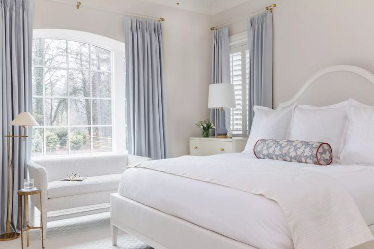 a white bed sitting in a bedroom next to two windows with blue drapes on them