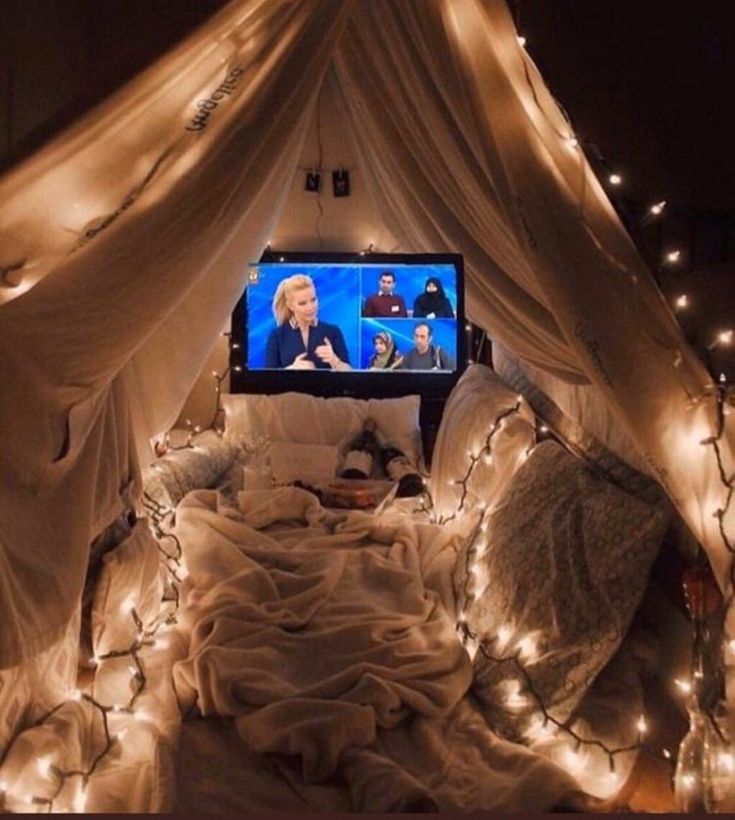 a bed covered in white sheets and lights with a tv on top of the bed