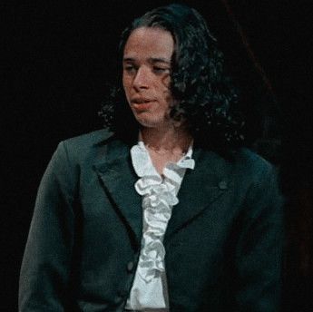 a man with long hair wearing a black jacket and white shirt standing in front of a dark background