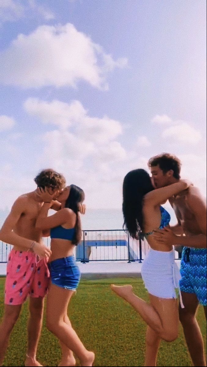 four people in bathing suits are standing on the grass by the water and one person is holding his arm around another man's neck