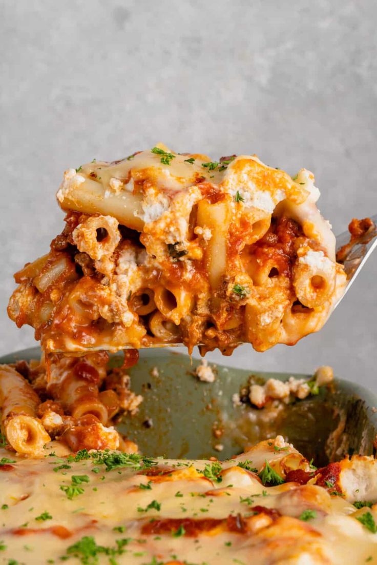 a spoon full of pasta being lifted from a casserole dish with cheese and sauce