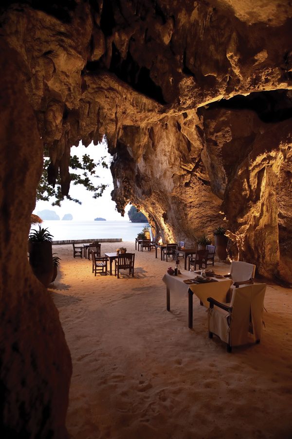 an outdoor dining area with tables and chairs on the beach next to a cave like structure