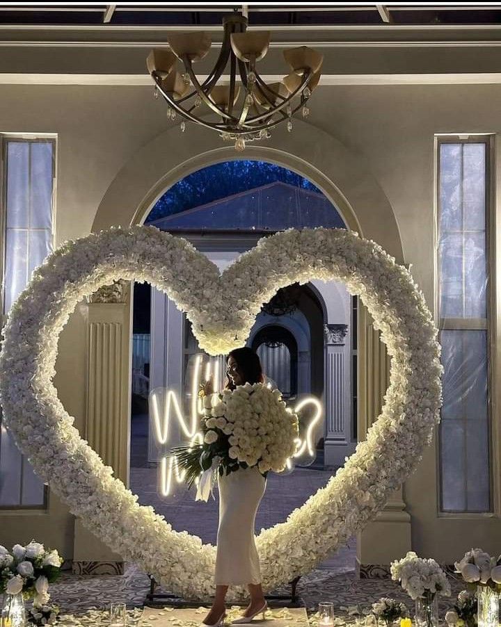 a woman standing in front of a heart shaped archway with flowers on the floor and lights behind her