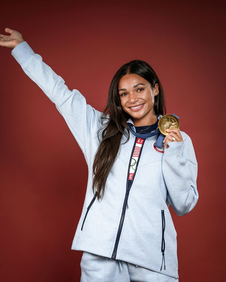 a woman is holding her gold medal in one hand and smiling at the camera while standing against a red background