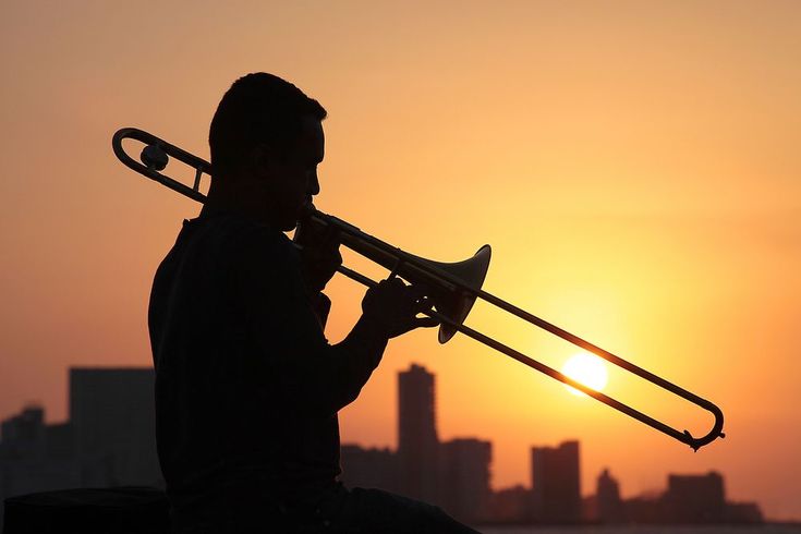 a man holding a trumpet while standing in front of the sun with buildings behind him