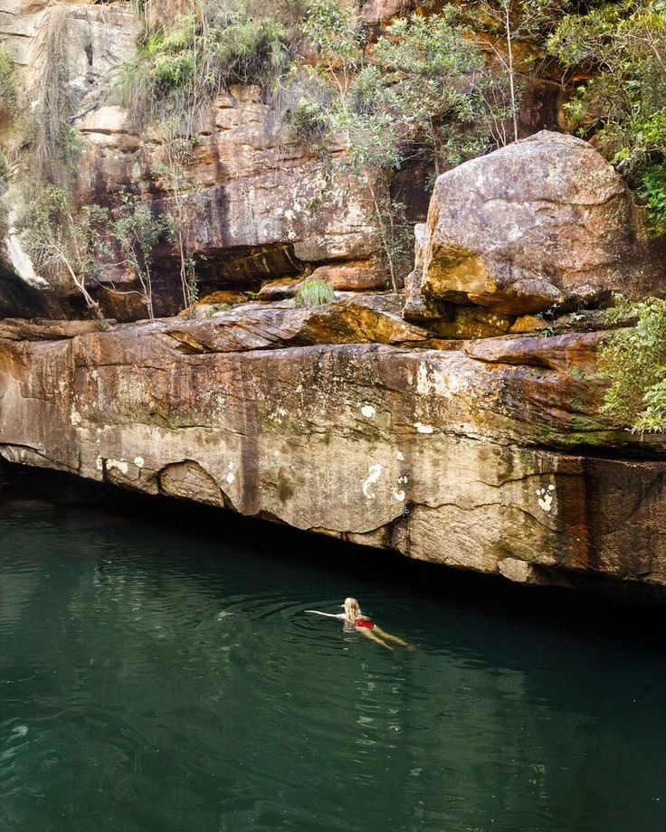 a person swimming in the water near some large rocks and trees on either side of a cliff