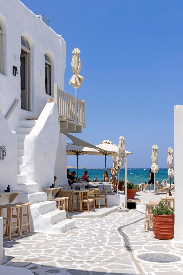 an outdoor dining area with tables and umbrellas on the beach, next to white buildings