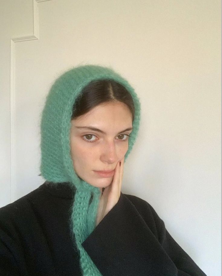 a woman wearing a green knitted hoodie and black jacket with her hand on her chin